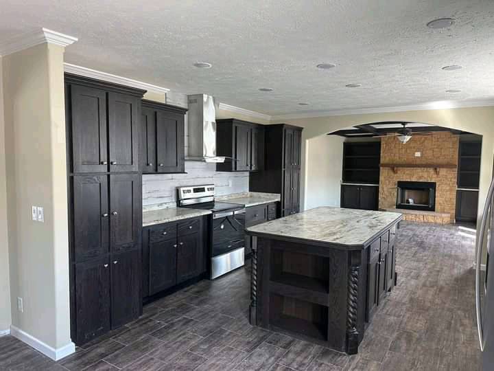 Spacious kitchen with dark wood cabinets, central island, stainless steel appliances, and adjacent living area featuring a brick fireplace. Cozy atmosphere.
