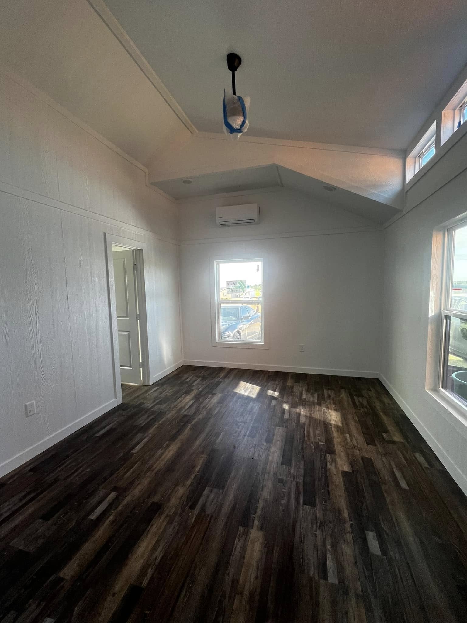 Spacious room with high ceilings, dark wood flooring, and white walls. A window lets in natural light. An air conditioner is mounted high on the wall.