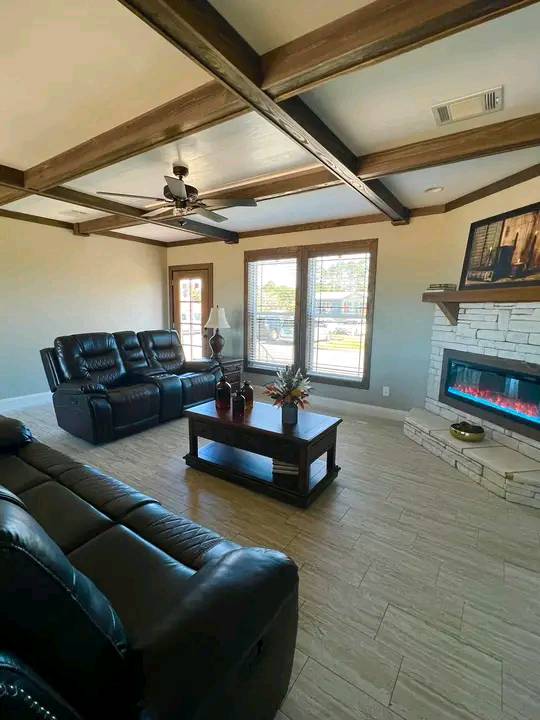 Living room with black leather sofas, wooden coffee table, and electric fireplace. Exposed beams on ceiling add warmth, large windows bring in light.