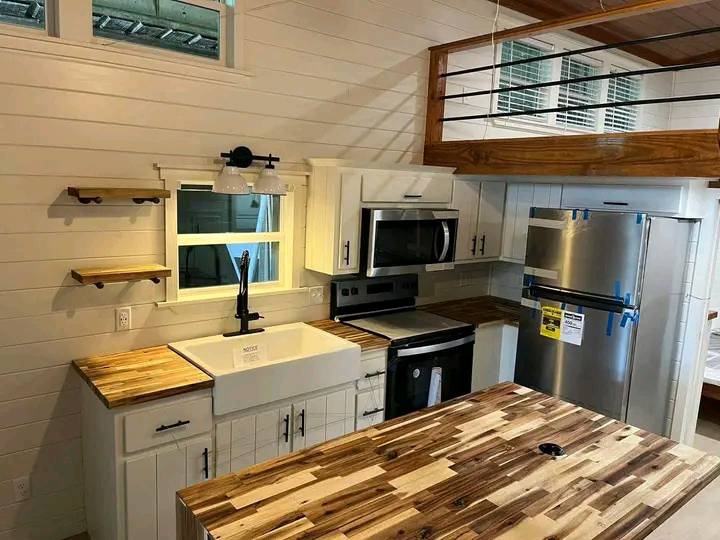Modern kitchen with white cabinets, wood countertops, and stainless steel appliances. Features a farmhouse sink, wall shelves, and a loft area above. Cozy and rustic.