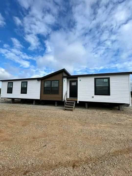 Single-story prefab home with white siding, dark window frames, and a central brown section. Set on a gravel lot under a partly cloudy sky.