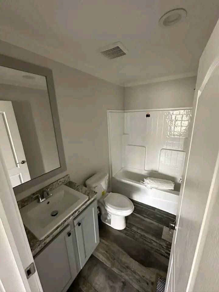 Small, modern bathroom with gray and white tones. Features a sink with a large mirror, toilet, and a shower-tub combo. The atmosphere is clean and minimalistic.