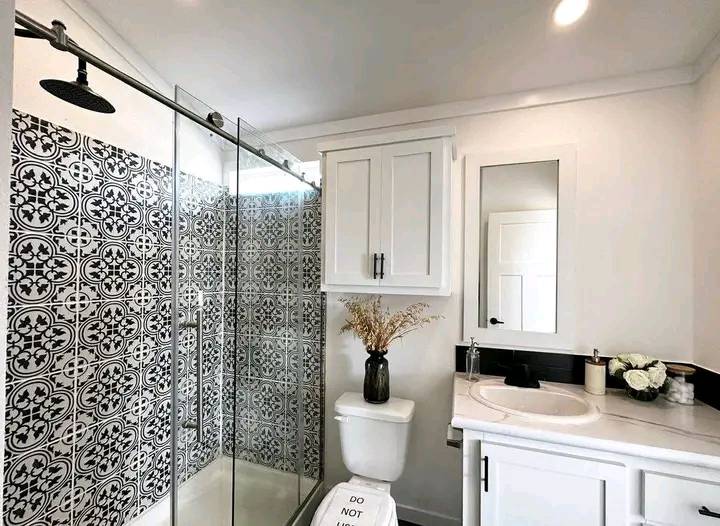 A modern bathroom with a white and black theme. Features include a shower with patterned tiles, a vase of dried flowers on the toilet, and a vanity with a mirror.