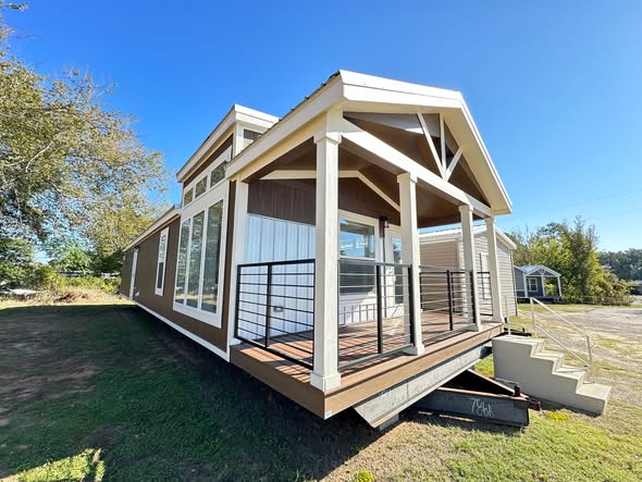 A modern, brown and white tiny house with large windows and a front porch. The porch features a metal railing, and steps lead up to it. The setting is bright and sunny.