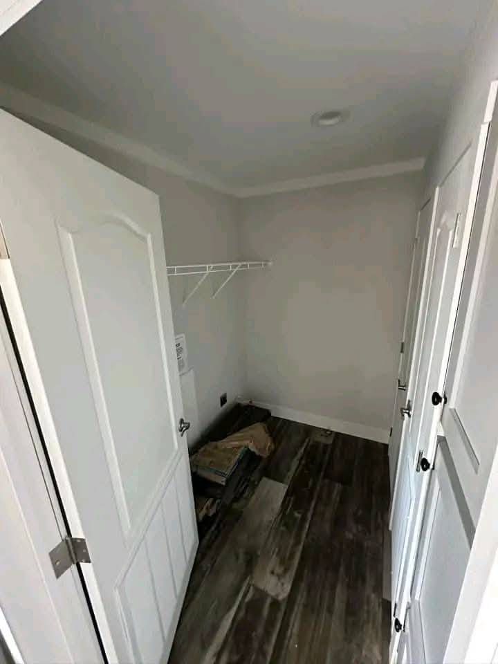 Small, empty room with white walls and an open door on the left. A wire shelf is mounted on the wall, and the wooden floor has a few items stacked in the corner.