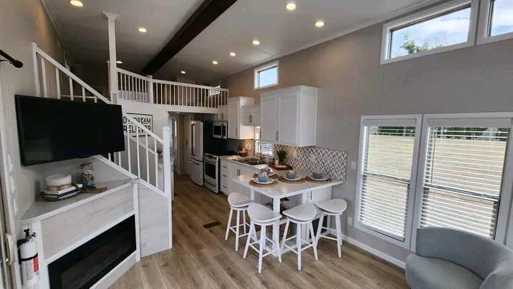 Modern, cozy tiny home interior with a loft. Features a kitchen with white cabinets and bar stools, a fireplace, and a wall-mounted TV. Bright, airy ambiance.