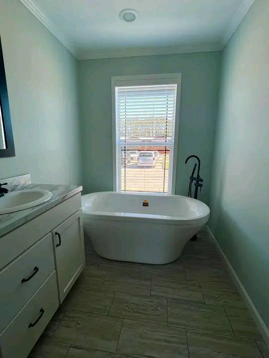 Minimalist bathroom with a white freestanding tub under a window with blinds. Light green walls and natural tones create a calm atmosphere.