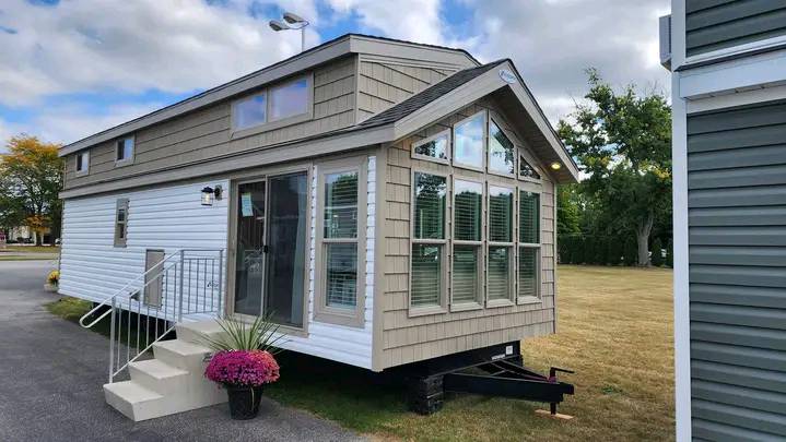 A modern tiny house on wheels with tan and white siding sits on a driveway. Large windows, front steps, and a pink flower pot create a welcoming look.