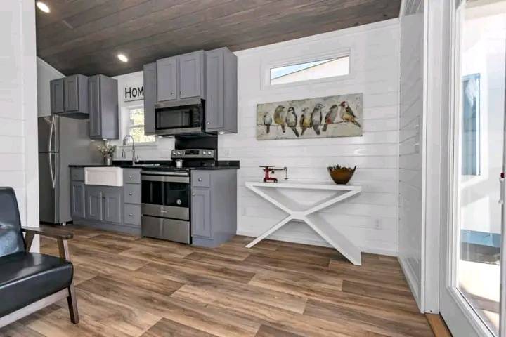 Modern kitchen with gray cabinets, stainless steel appliances, and a farmhouse sink. Bright space with wooden floor and bird art on the wall. Cozy ambiance.