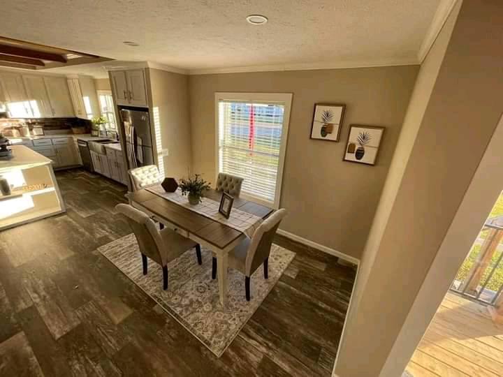 Bright dining area with a wooden table, four cushioned chairs, and a decorative plant centerpiece. Warm sunlight filters through blinds, illuminating the room’s earthy tones and wooden floor. Two framed botanical prints adorn the beige walls, creating a cozy, inviting ambiance.