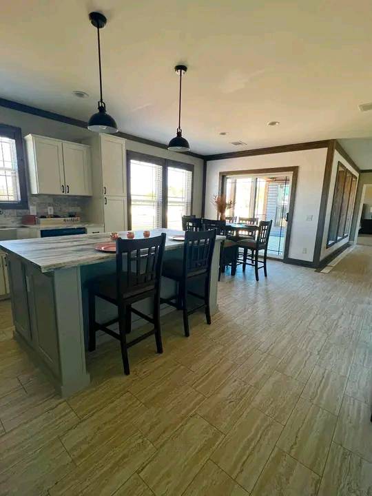 Spacious kitchen with a marble island, high-backed chairs, pendant lights, and wood cabinets. Dining table by large windows, creating a cozy, bright atmosphere.