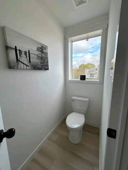 A small, bright bathroom with white walls and a wooden floor. It features a toilet beneath a window showing a cloudy sky. A black-and-white pier photo hangs on the left wall.