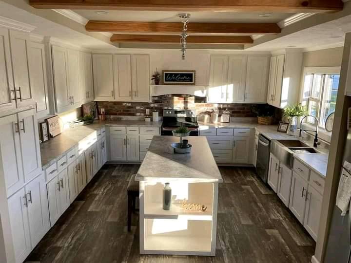 Bright, spacious kitchen with white cabinets, marble island, and wood beams. Sunlight streams through windows, creating a warm, welcoming atmosphere.
