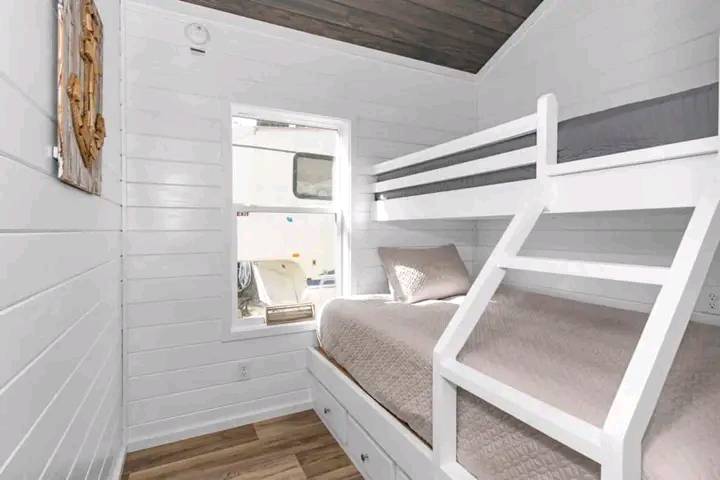 A cozy bedroom with white shiplap walls features a bunk bed with a gray quilt. Sunlight streams through a window, illuminating the wooden floor.
