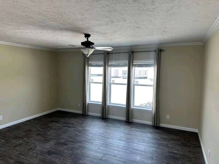 Spacious, empty room with beige walls, dark wood flooring, and three large windows with gray curtains. A ceiling fan hangs from the textured ceiling.
