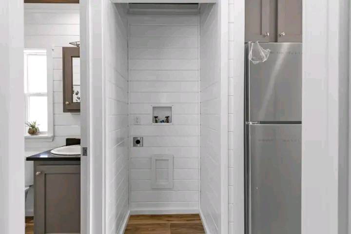 Narrow laundry nook with white shiplap walls, empty space for washer and dryer. Adjacent kitchen area visible with fridge and cabinets, creating a clean, modern look.