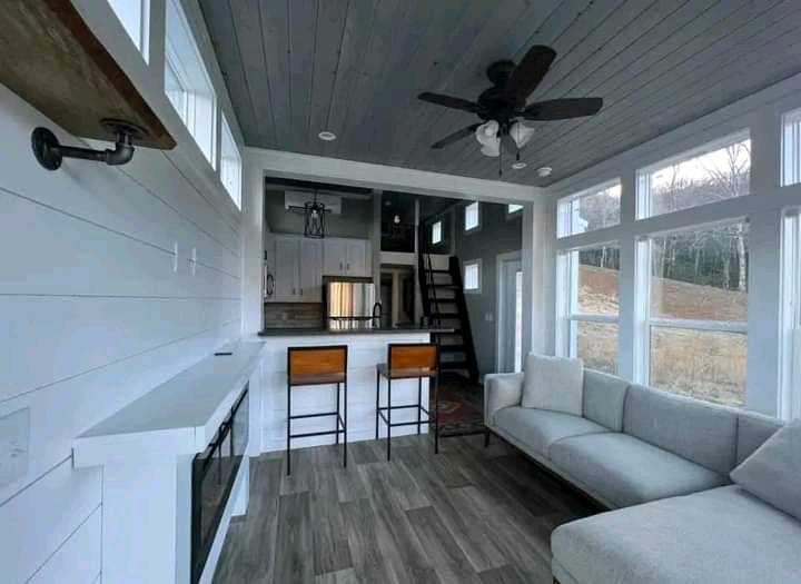 A modern tiny house interior with white walls, large windows, and wood flooring. Features a sleek kitchen, high ceiling, cozy gray sofa, and loft area.
