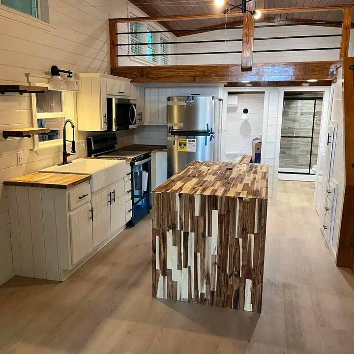Modern kitchen with white cabinets, stainless steel appliances, and a large wooden island with varied grain patterns. Loft area visible above. Cozy and warm ambiance.
