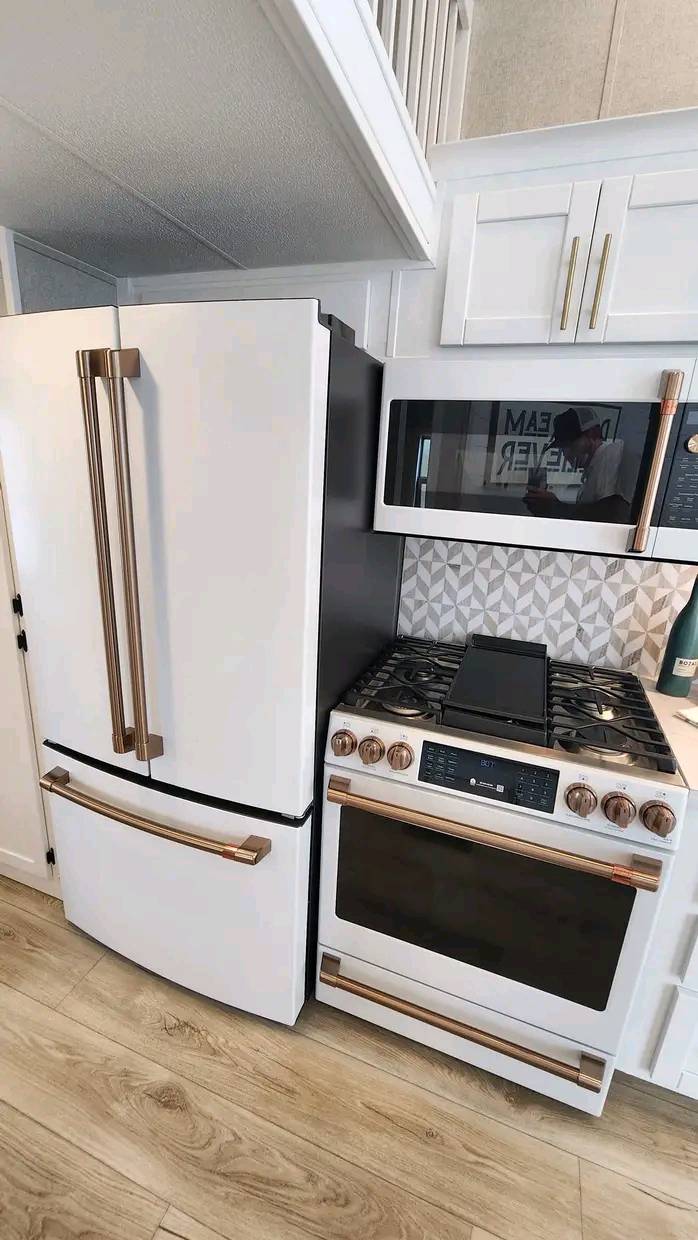 Modern kitchen featuring a white refrigerator with bronze handles beside a matching oven and stove. Geometric tile backsplash, light wood floor.