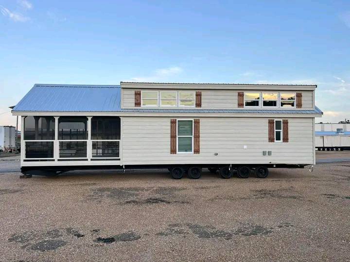 A beige mobile home on wheels with a blue metal roof sits on gravel. It has a screened porch, multiple windows, and a calm, open sky above.