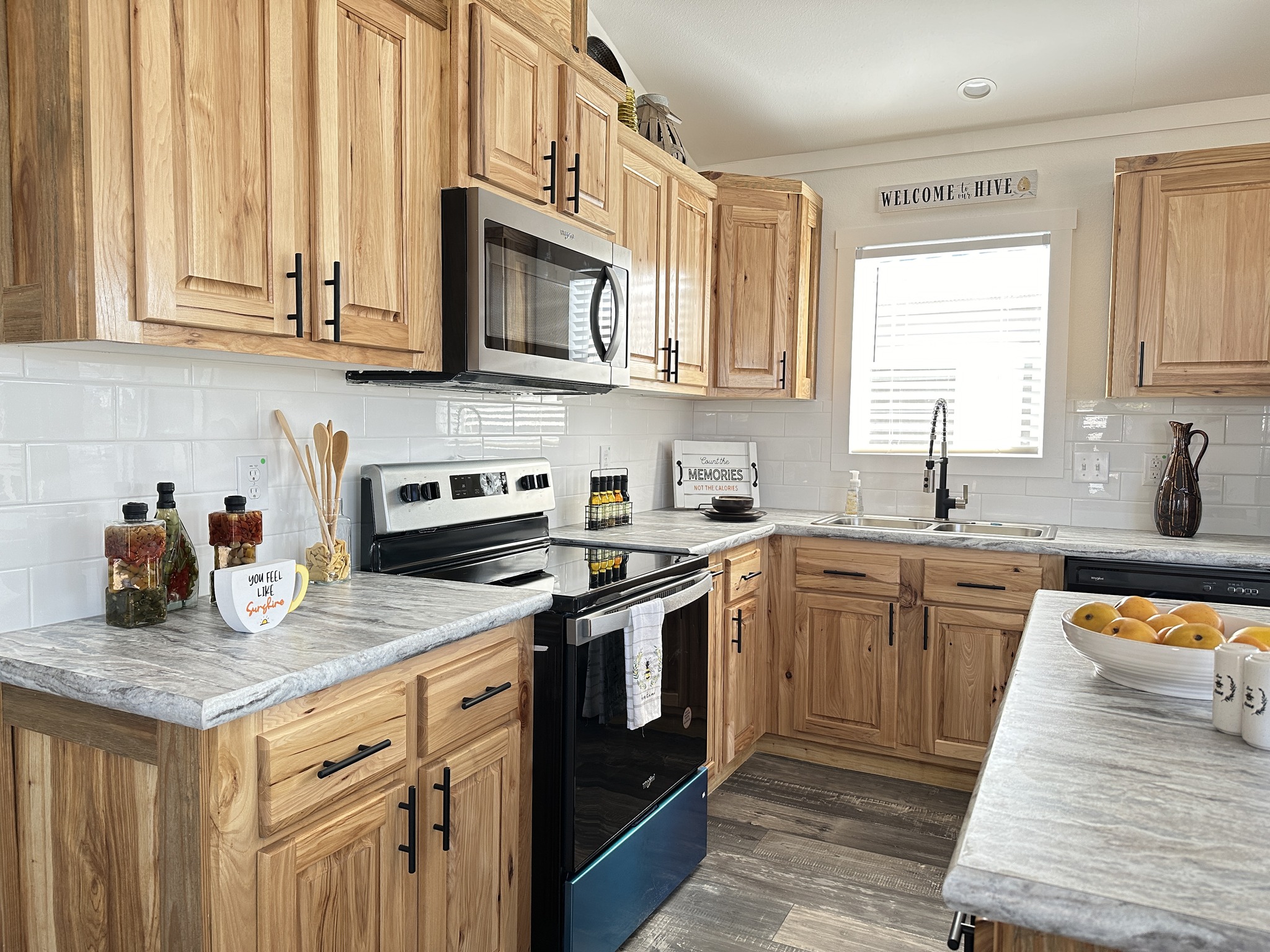 Warm kitchen with natural wood cabinets, granite countertops, and a stainless steel oven. Bright window, cheerful decor, and a bowl of oranges add freshness.