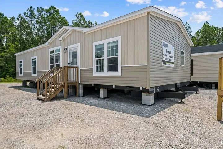 Tan modular home with white-trimmed windows, set on blocks in a gravel lot. Surrounded by trees under a blue sky, creating a serene and inviting scene.