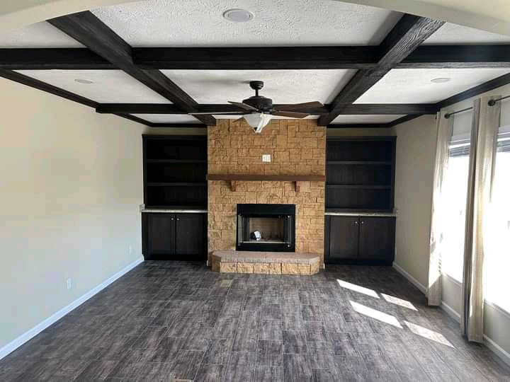 Spacious living room with a stone fireplace, dark wood ceiling beams, and shelves. Sunlight filters through curtained windows, creating a cozy ambiance.