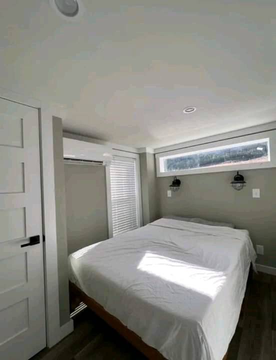 A small, cozy bedroom with a neatly made bed covered in a white sheet under a narrow horizontal window. Minimalist decor, soft natural light, and wall sconces create a serene atmosphere.