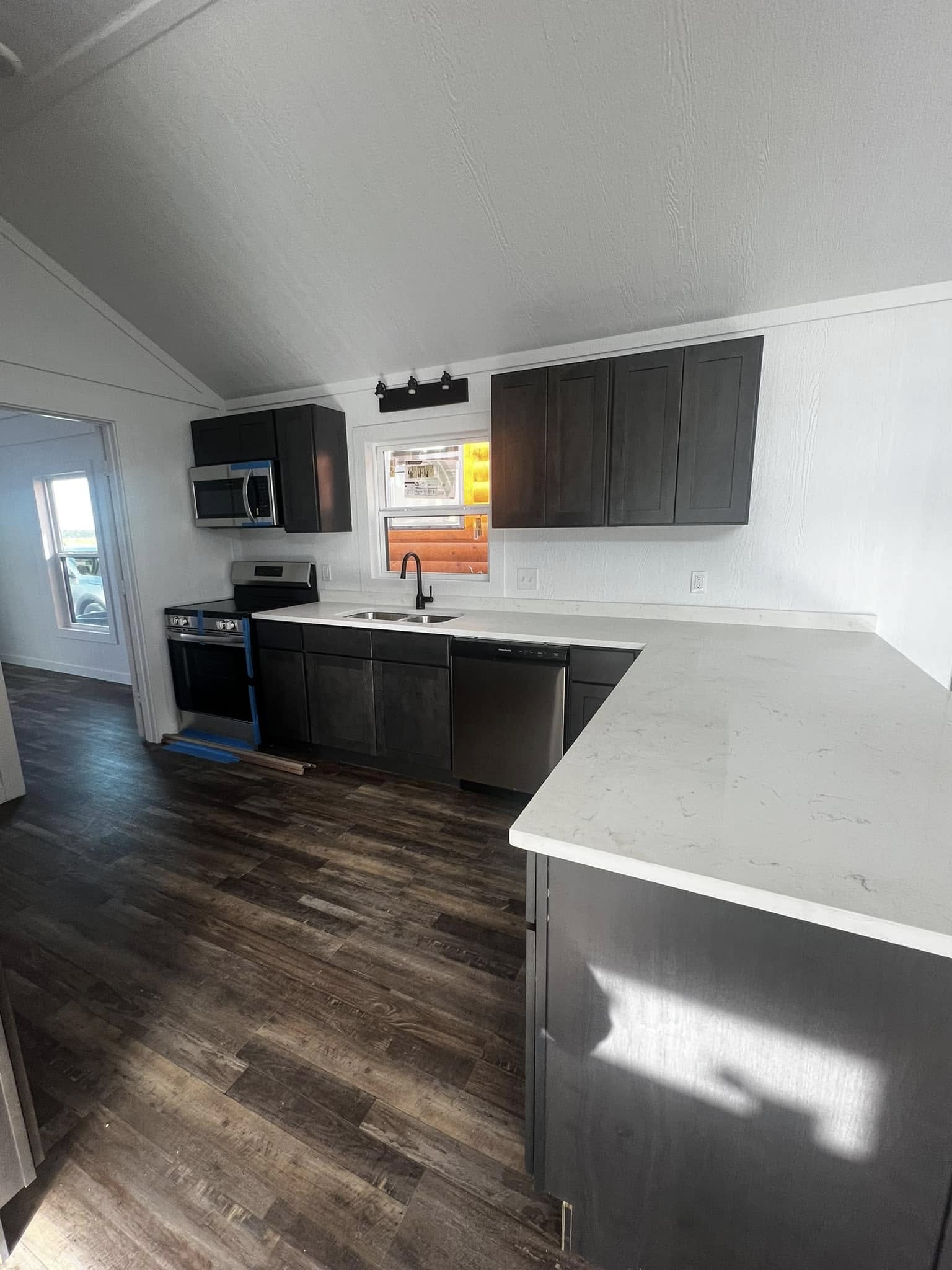 Modern kitchen with dark wood cabinets, white countertops, stainless steel appliances, and dark wood flooring. Sunlight streams in, creating a warm ambiance.