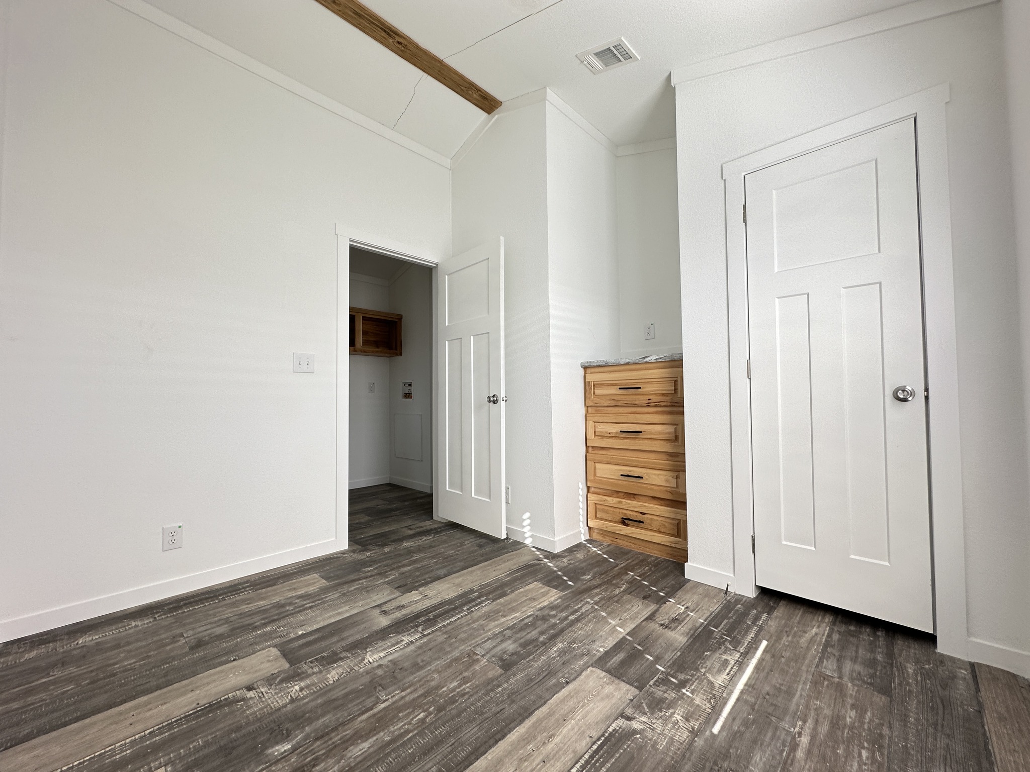 Bright, empty room with white walls and wood-look flooring. Features a wood dresser, two white closet doors, and a visible ceiling beam.
