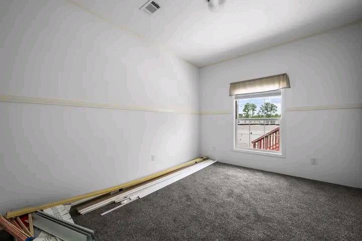 Empty room with white walls, gray carpet, and a single window with a beige valance. Construction materials are stacked on the floor.