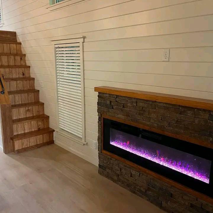 Cozy interior with a wooden staircase beside a modern stone fireplace emitting purple flames. White paneled walls add a warm, inviting feel.