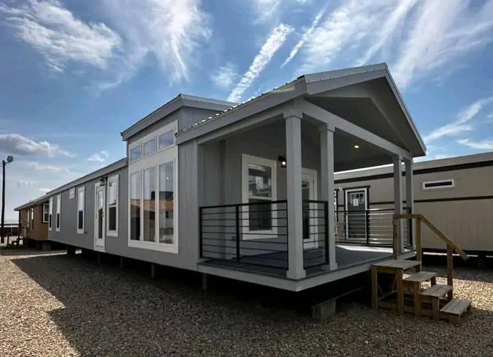 Modern tiny house with a gray exterior and large windows, set on gravel. A small porch with railings and steps is visible. The sky is partly cloudy.