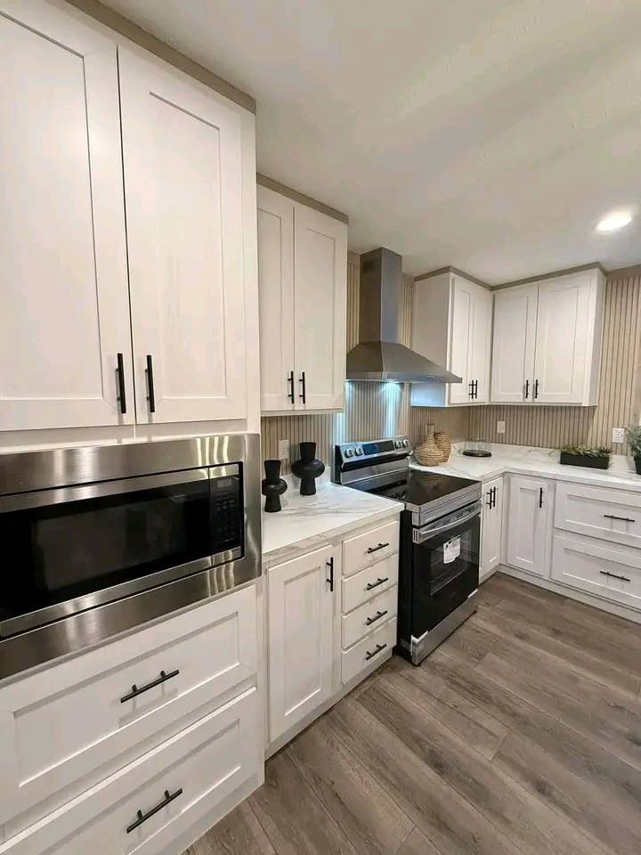 Modern kitchen with white cabinets, stainless steel appliances, and wood flooring. Features a microwave, oven, range hood, and elegant decor accents.