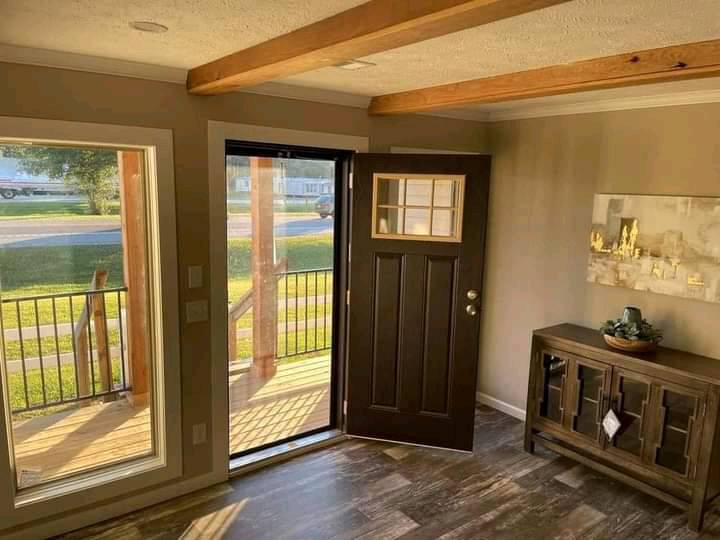 Cozy entryway with a dark wooden door ajar, sunlight streaming through large windows. Wooden beams on the ceiling add warmth. A console table with decor on the right.