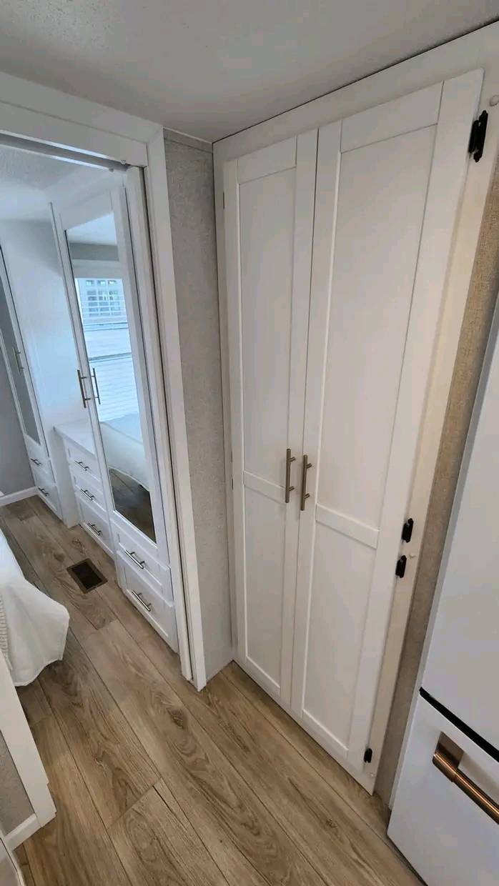 A narrow hallway with light wood flooring features white double cabinets on the right, a mirrored wardrobe with drawers on the left, and a white wall. Bright, minimalist, serene.