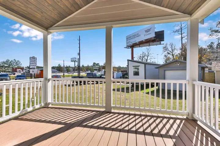 View from a wooden porch with white railing, overlooking a grassy area and road with parked cars. A billboard and trees are visible under a clear sky.