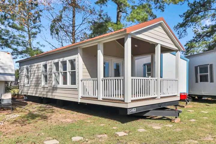 A small, cream-colored tiny house with a red roof sits on a grassy yard, surrounded by trees. The house features large windows and a welcoming porch.