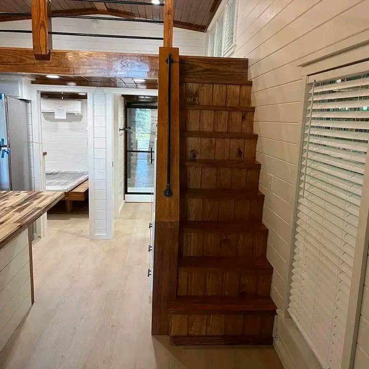 Wooden staircase with black rail leads to loft in a cozy, wooden-finished interior. Nearby, a kitchen counter and bedroom are visible, creating a warm, homey atmosphere.