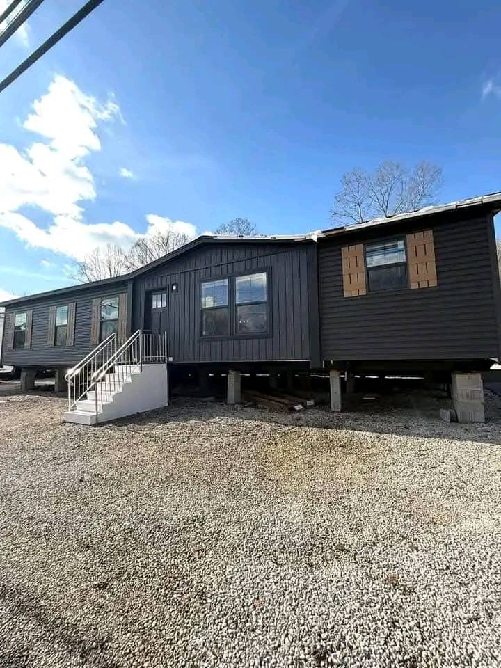 Dark brown, elevated mobile home with a white staircase, large windows, and wooden shutters. Situated on gravel under a clear blue sky, conveying calmness.