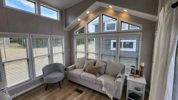 Cozy living room with large, bright windows, a light gray sofa adorned with pillows, and a striped throw. Soft armchair and side table enhance the inviting space.