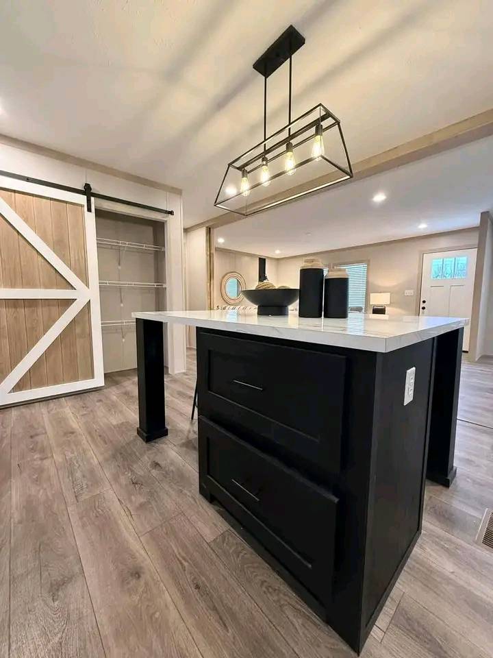 Modern kitchen interior with a central island featuring a black base and white countertop. Overhead, a stylish rectangular light fixture hangs. To the left, a sliding barn door opens to a closet with shelving. Wood-style flooring adds warmth.