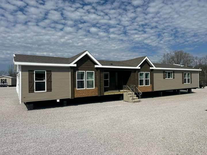 A large modular home with a two-tone exterior of beige and dark brown sits on a gravel lot under a partly cloudy sky. Steps lead to the main entrance.