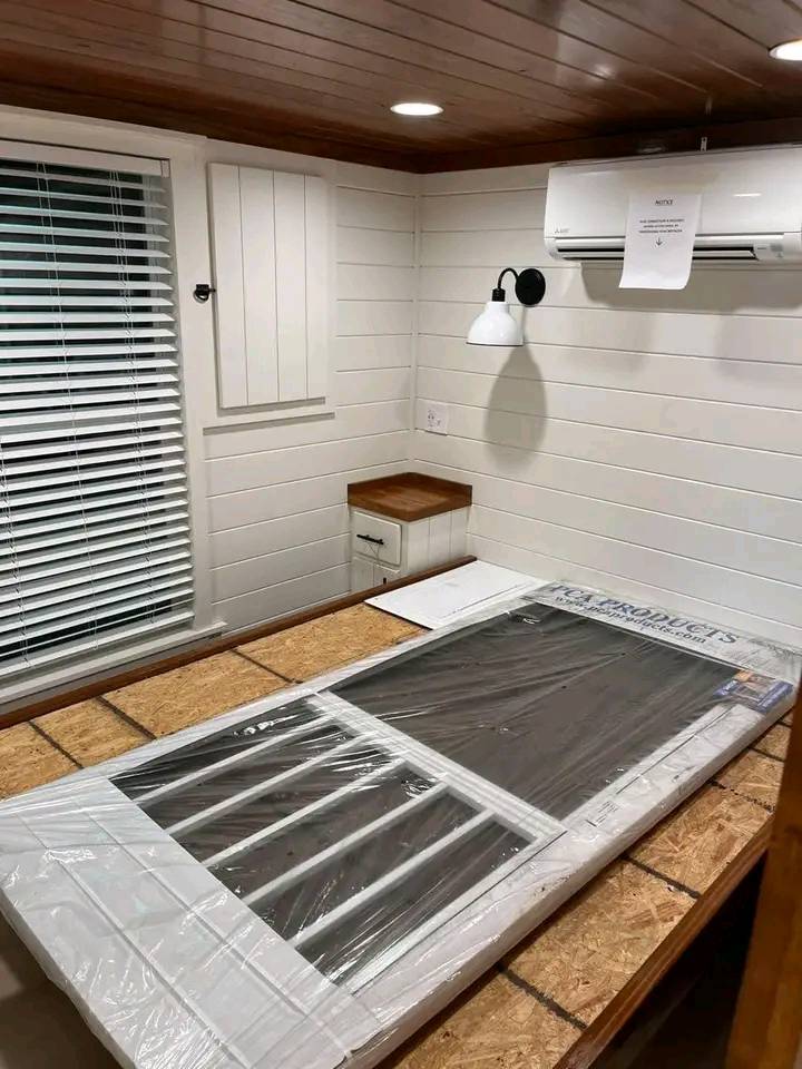 Cozy wooden room with white paneled walls, a window with blinds, and a ceiling-mounted air conditioner. A lamp and a sealed package lie on an unfinished bed frame, creating an intimate, rustic atmosphere.