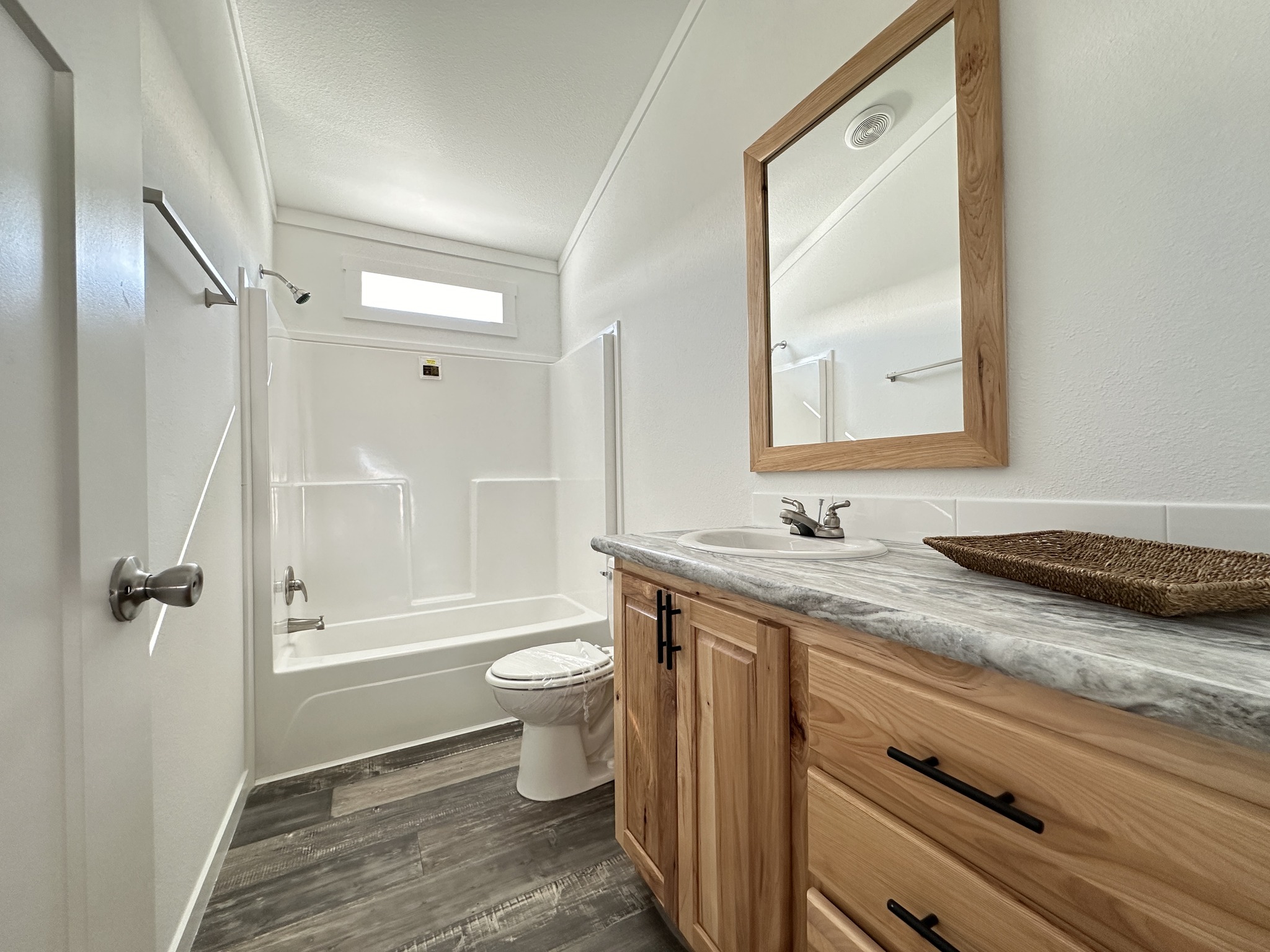 Modern bathroom with a wooden vanity, marble countertop, and large mirror. Bathtub and toilet are white, and the flooring is dark wood. Bright and clean.
