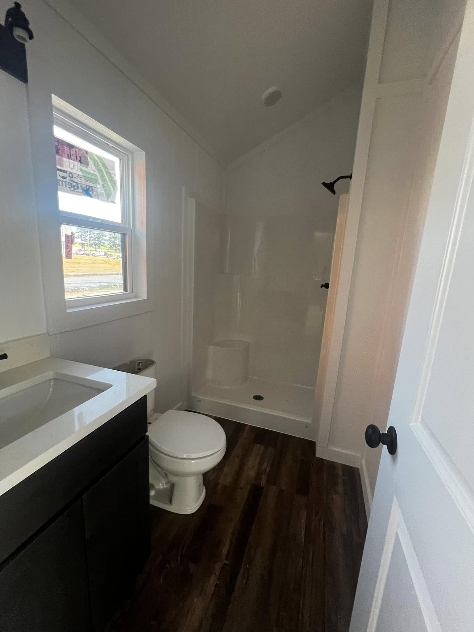Compact bathroom with a white shower, dark vanity, and wood floor. Natural light enters through a window, creating a fresh, clean atmosphere.
