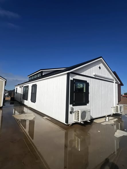 A modern, white prefab home with black window shutters sits on a wet surface under a clear blue sky, exuding a sleek, minimalist vibe.