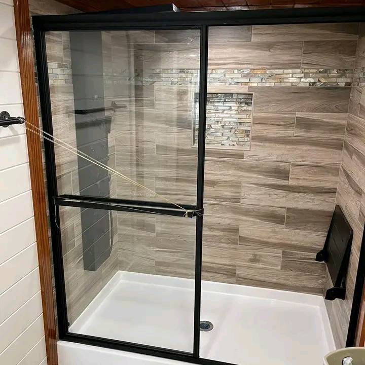 Shower with glass doors, black frame, and beige wood-patterned tiles. Decorative horizontal mosaic strip and a small inset shelf create a modern look.
