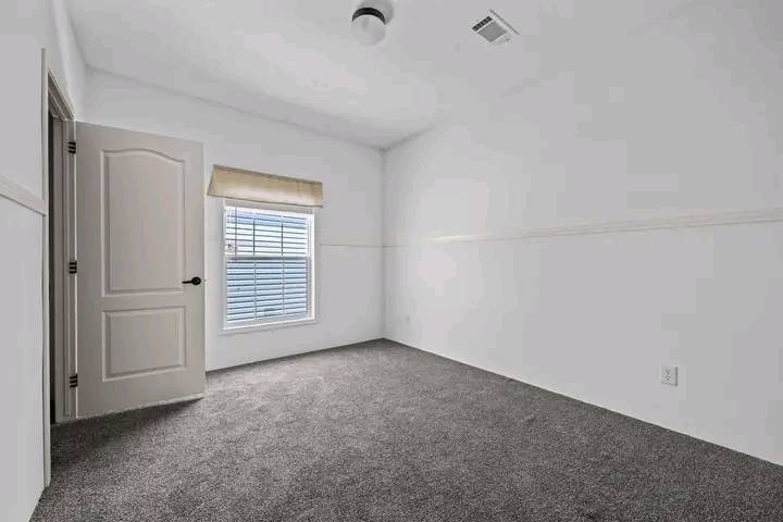 Small, empty room with white walls, gray carpet, and a single window with blinds. A closed white door stands on the left. The space feels quiet.