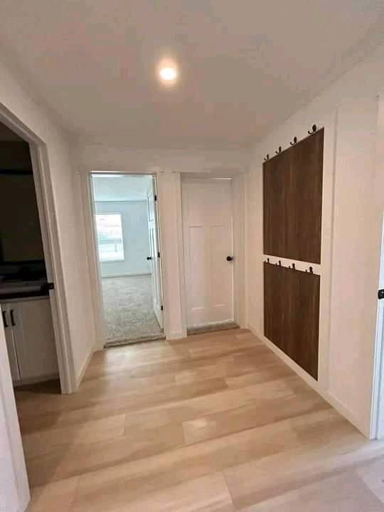 A hallway with light wood flooring leads to two white doors, one open to a room with carpet and a window. Dark wood paneling on the right wall. Bright and clean atmosphere.